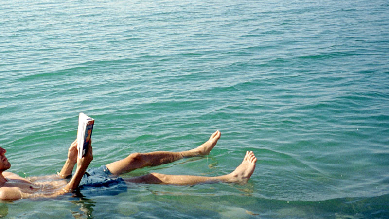 Man effortlessly floating on the Dead Sea's mineral-rich waters in Jordan, reading a book under the clear blue sky